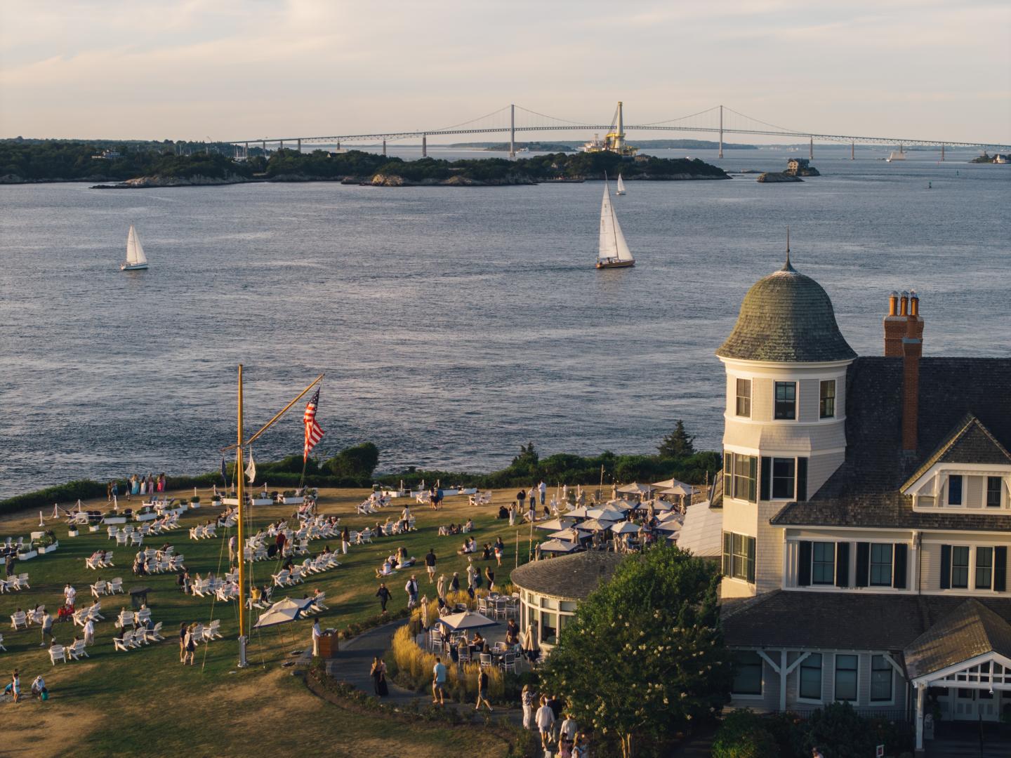 Coastal scene with sailboats, a grand house, and people gathered on a lawn.