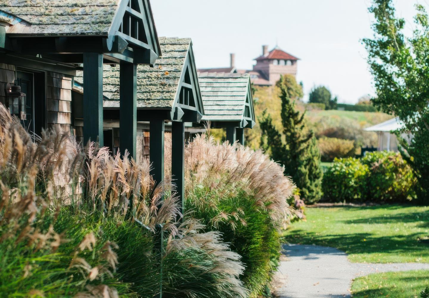Rustic cottages with greenery, pathway, and distant building in a sunny setting.