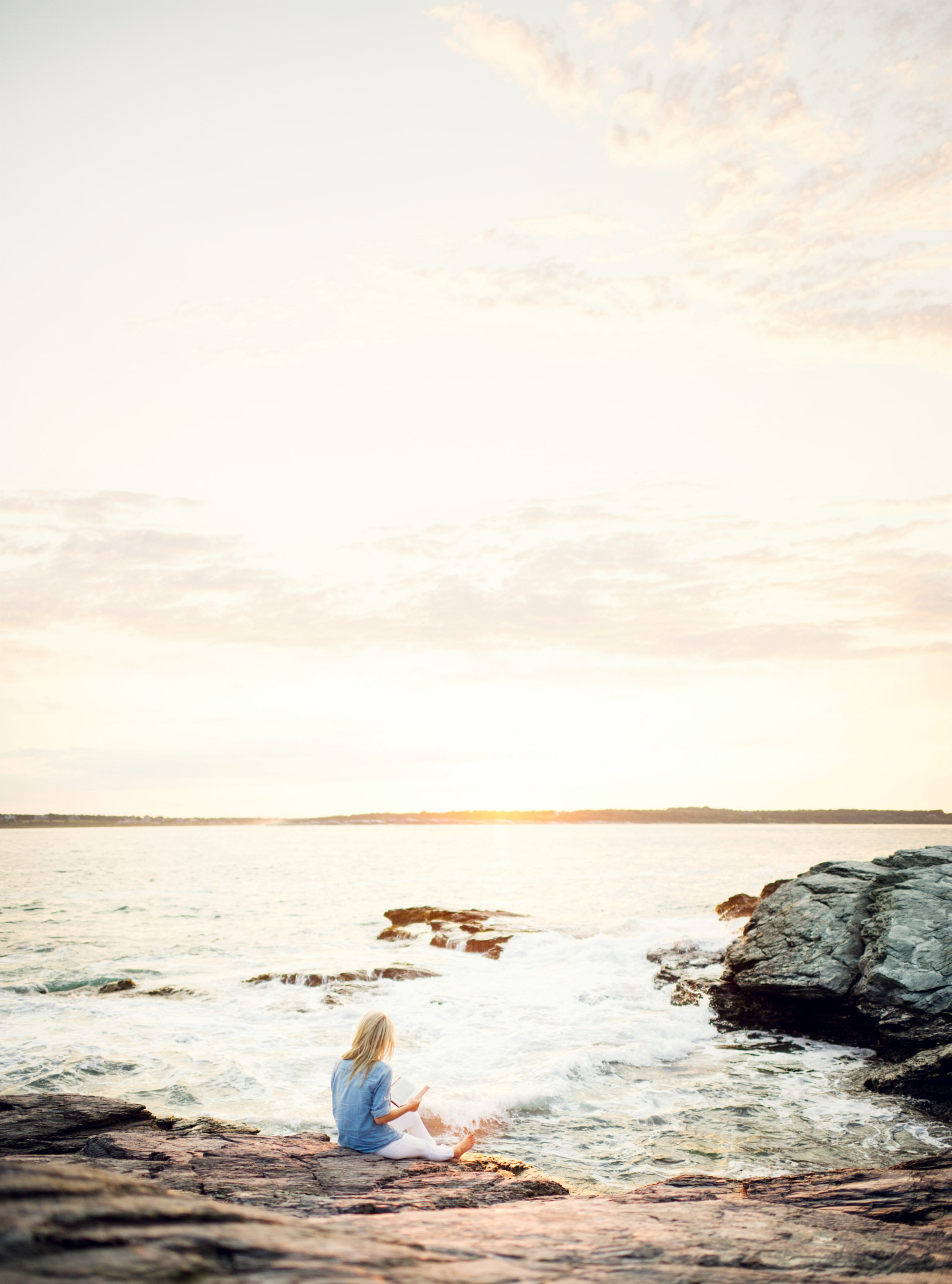 Seated person on rocky shore at sunset, with calm sea and cloudy sky.