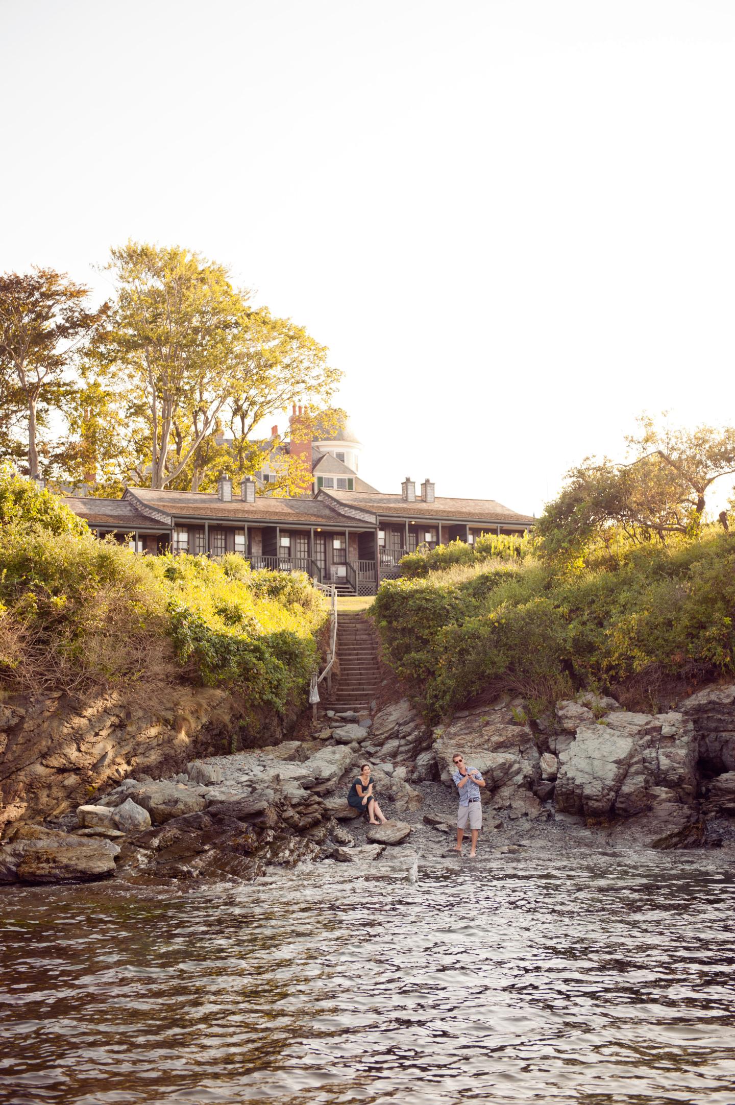 Rustic house atop a rocky shore with people by the water.
