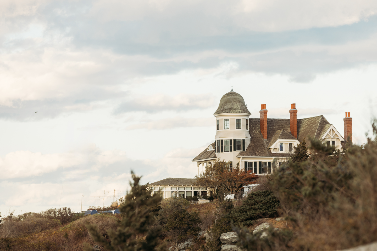 Coastal mansion with a turret, surrounded by trees under a cloudy sky.