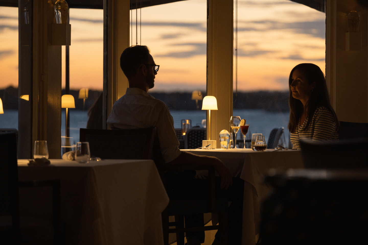 Couple dining at sunset by a window with a waterfront view.