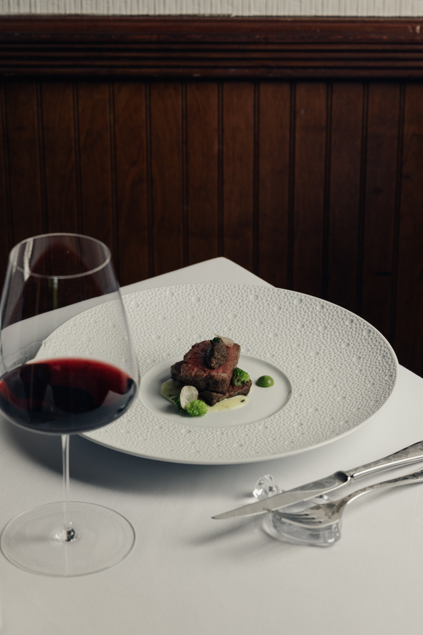 Seared meat on a white plate, with red wine and silverware on a white tablecloth.