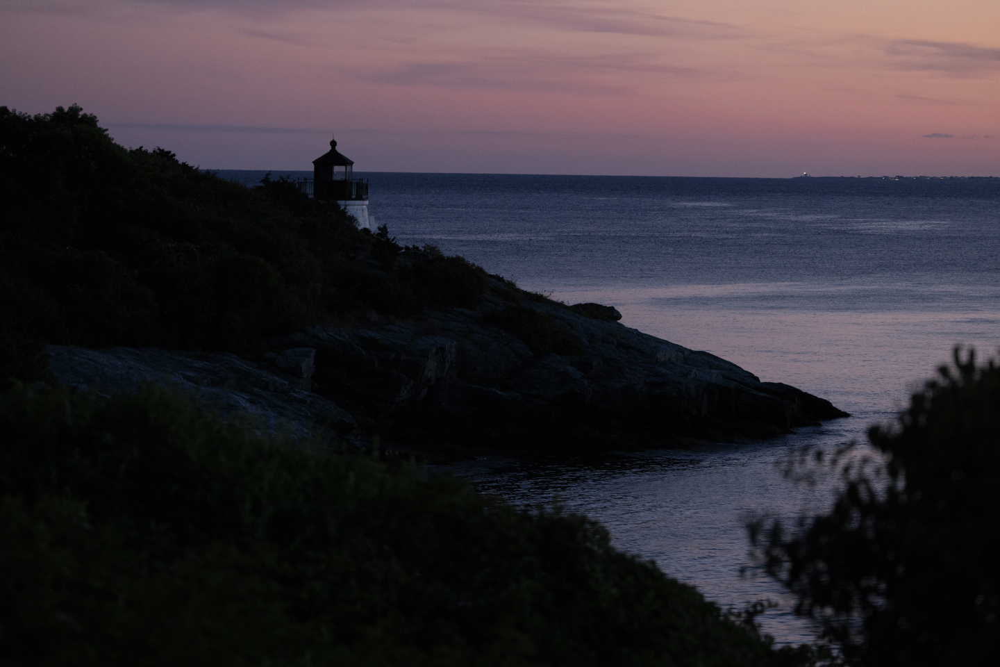 Coastal lighthouse at sunset with calm sea and dark silhouette of trees.