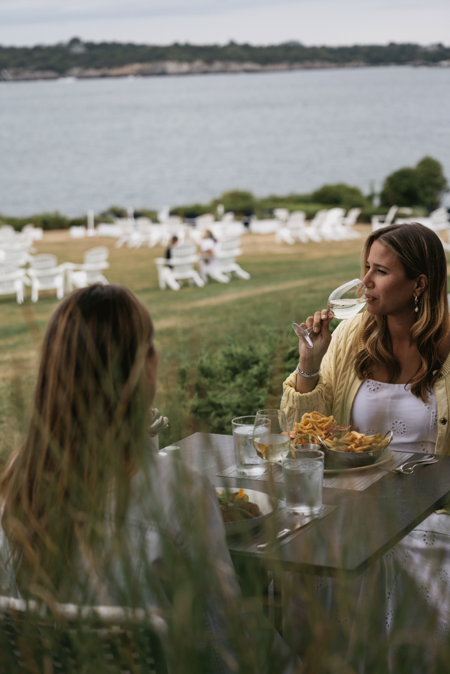 Two people dining outdoors by a lake, with white chairs on the grass.