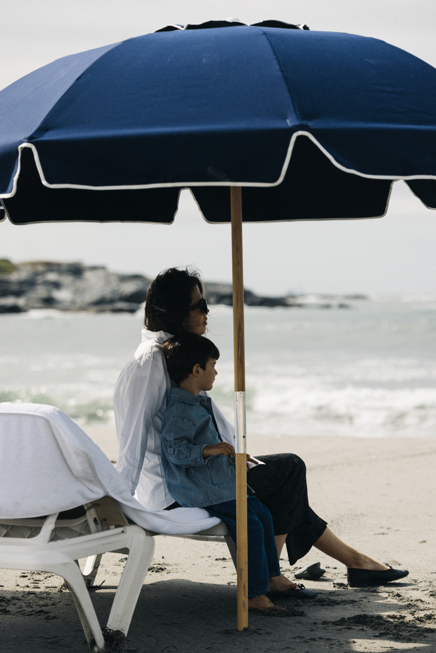 Woman and child sit under a blue beach umbrella by the ocean.