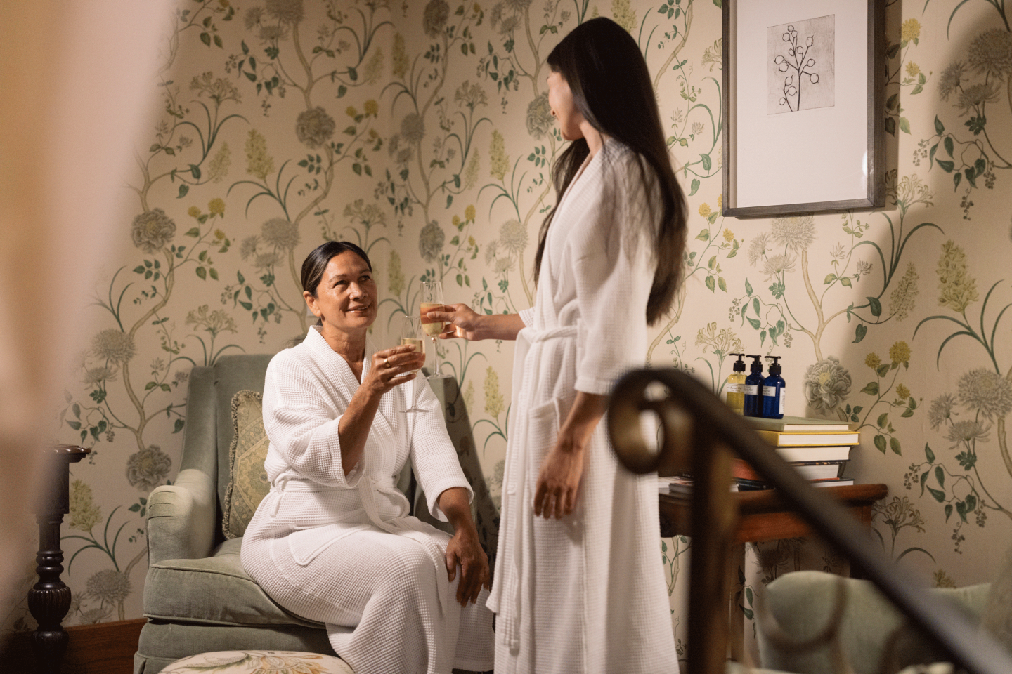 Two women in bathrobes enjoying drinks in a cozy, floral wallpapered room.