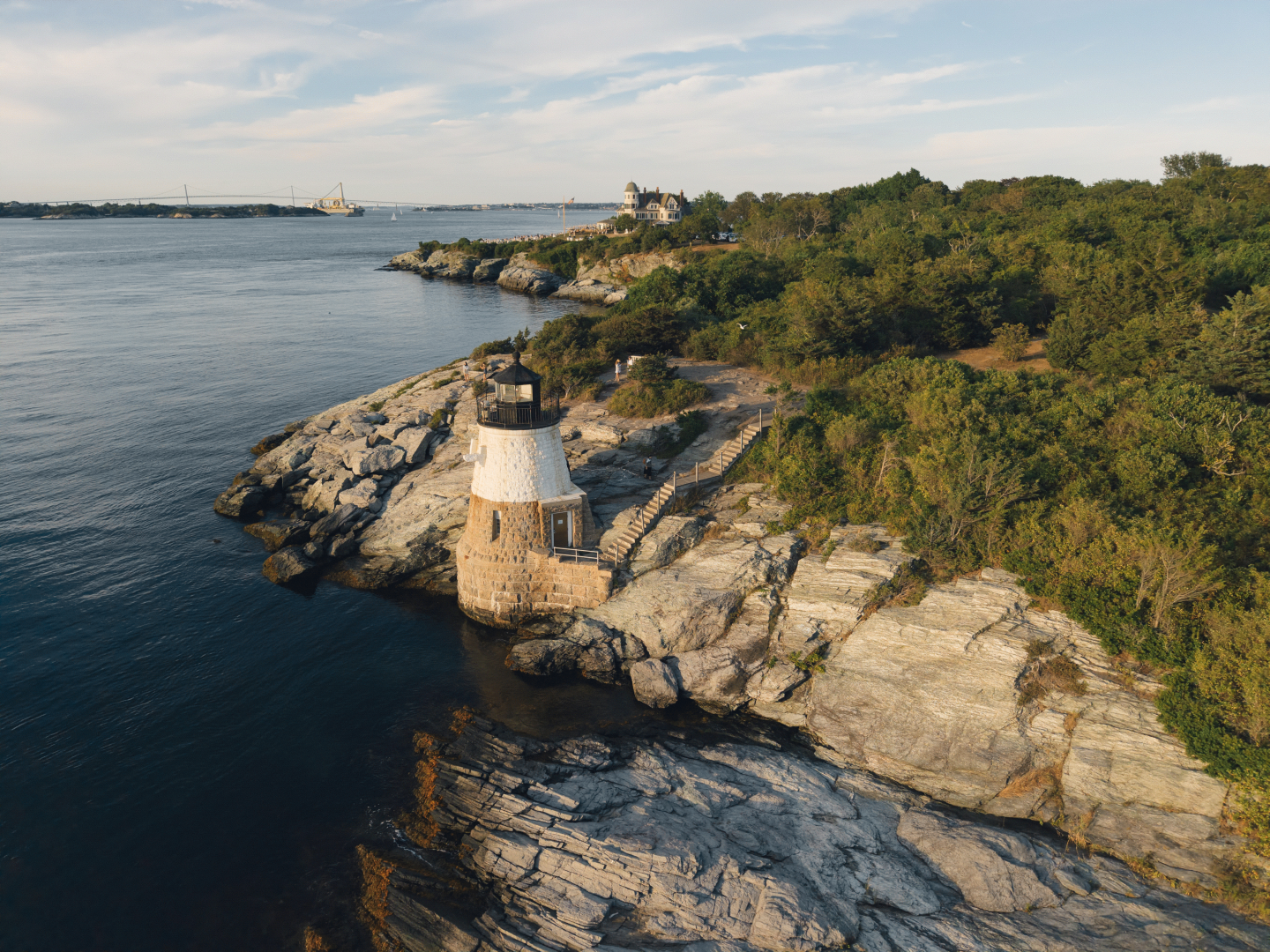 Lighthouse on rocky coastline with greenery and calm sea.