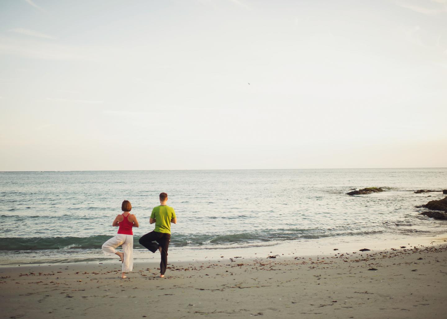 Two people practicing yoga by a calm ocean at sunset.