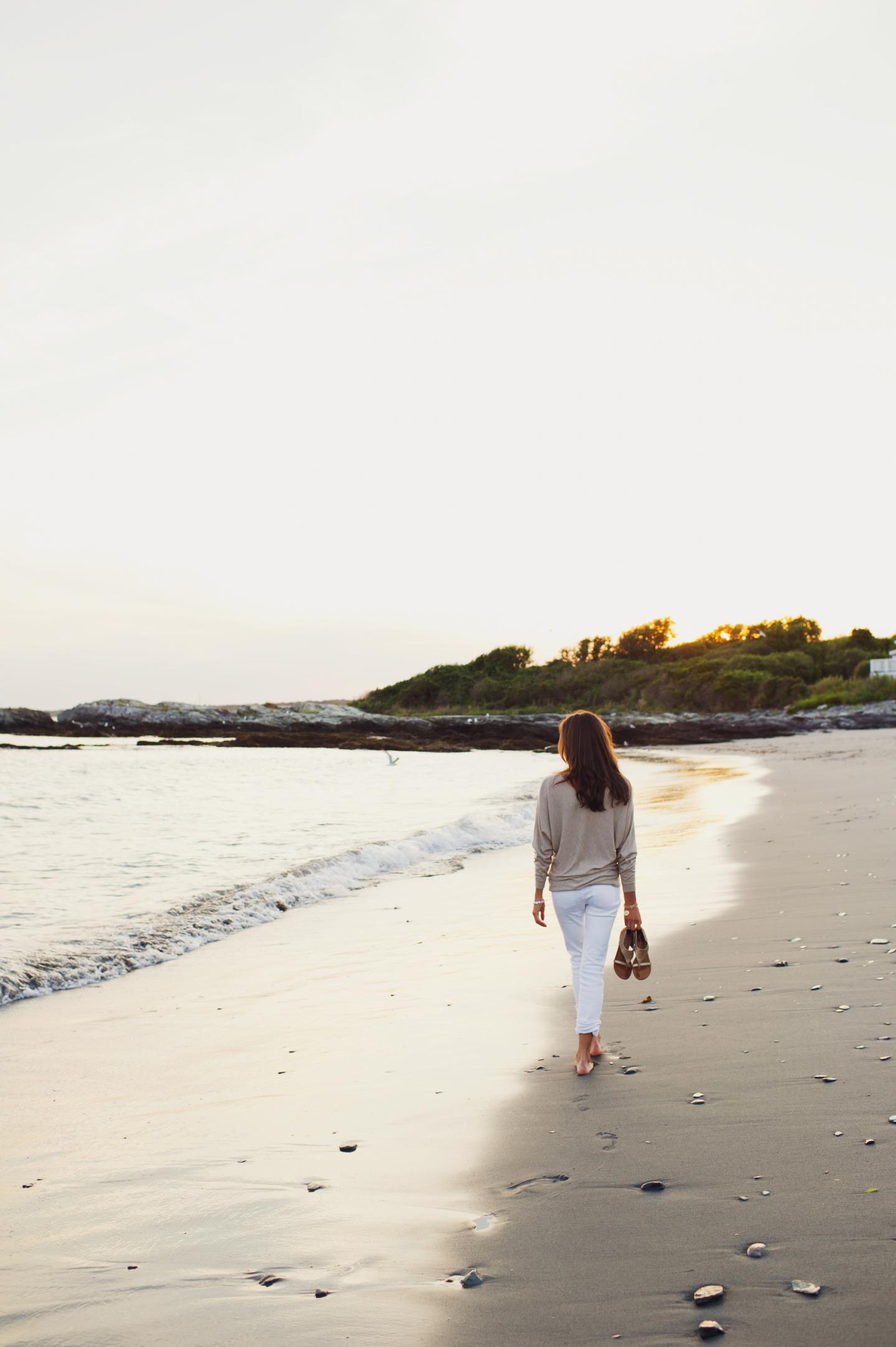 Woman walking on a sandy beach at sunset, holding sandals.