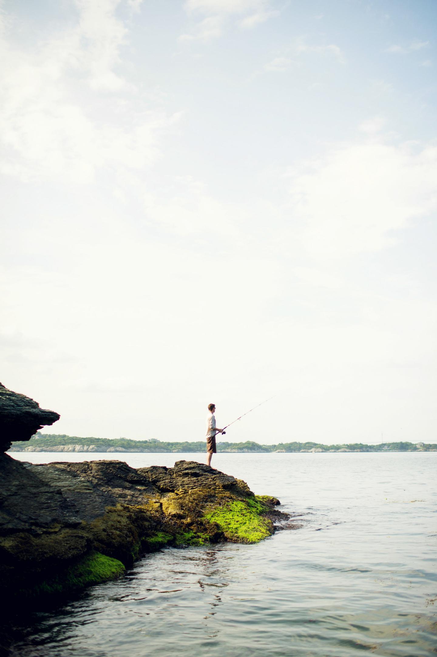 Person fishing on rocky shore under a cloudy sky.