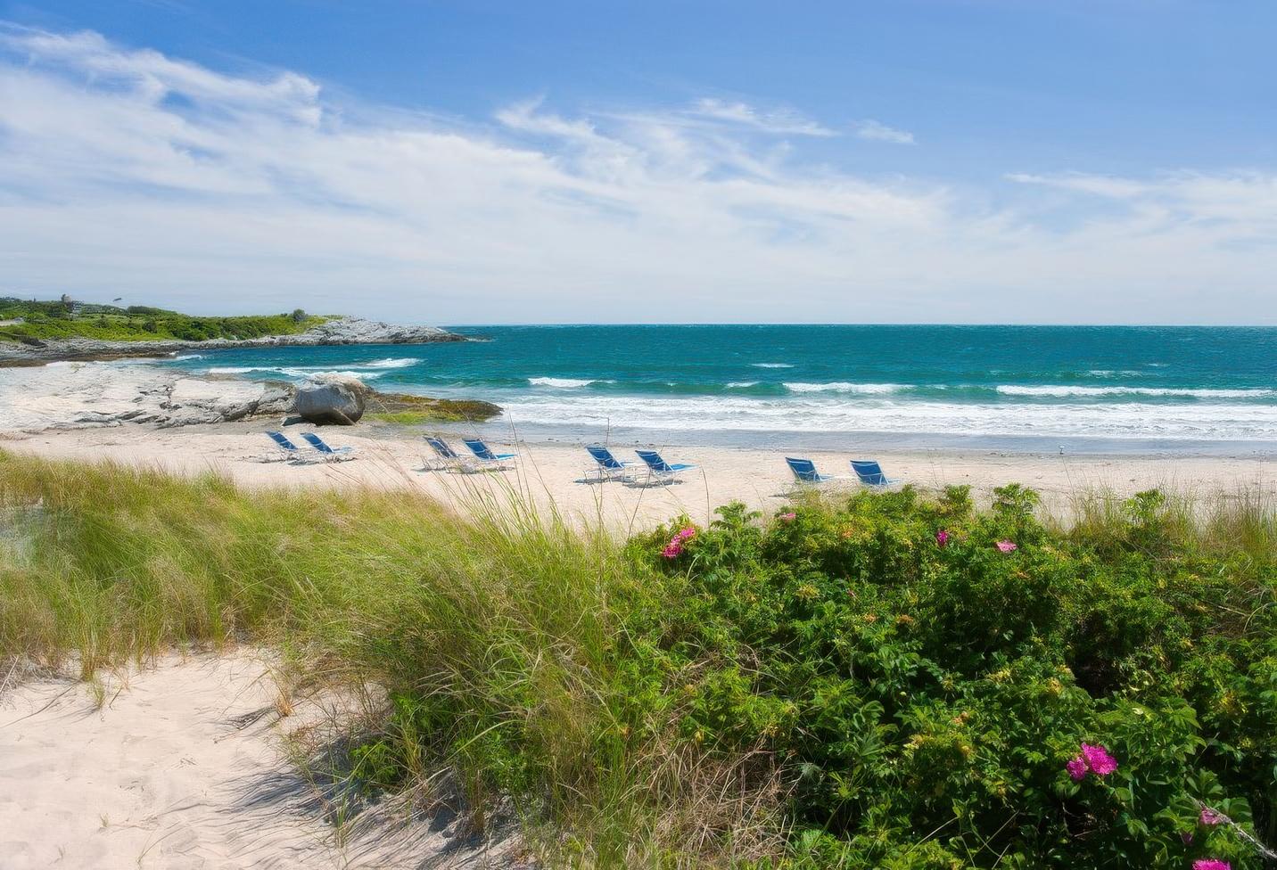 Sandy beach with lounge chairs, ocean waves, and grassy foreground.
