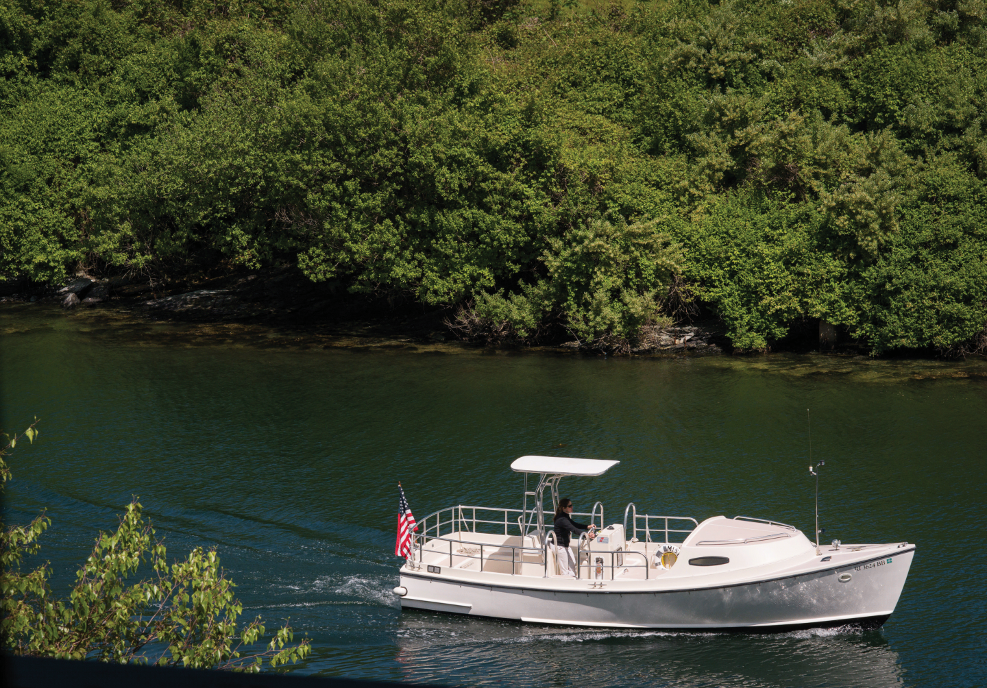White boat cruising on a green river beside lush trees.