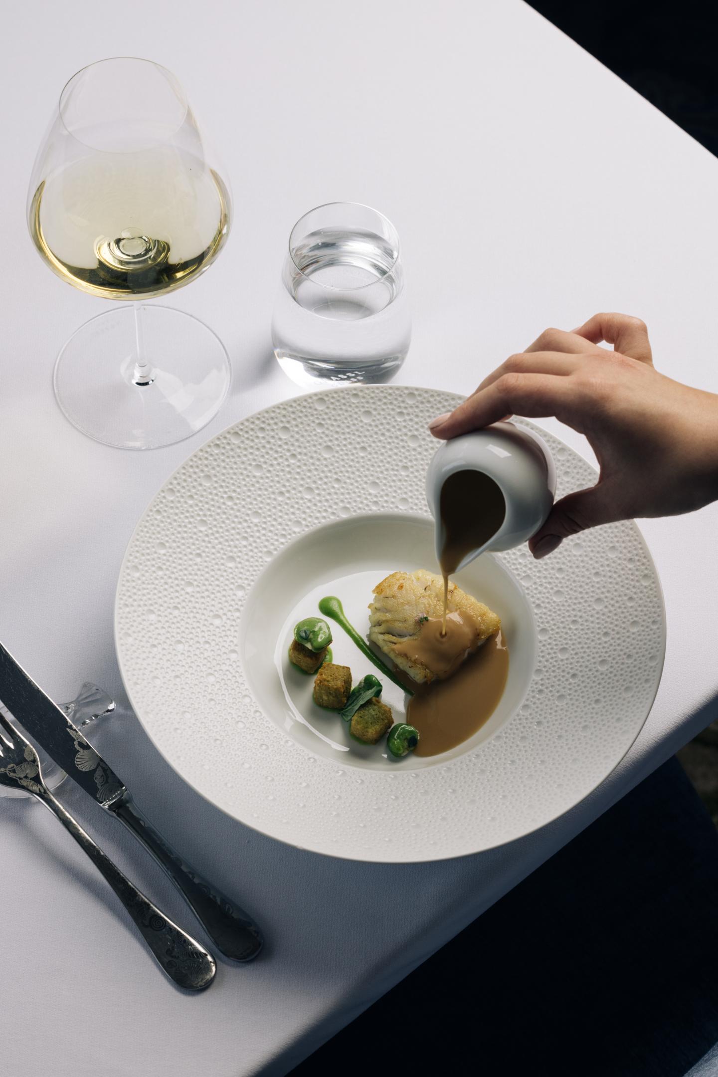 Gourmet dish with sauce poured, wine glass and cutlery on white tablecloth.