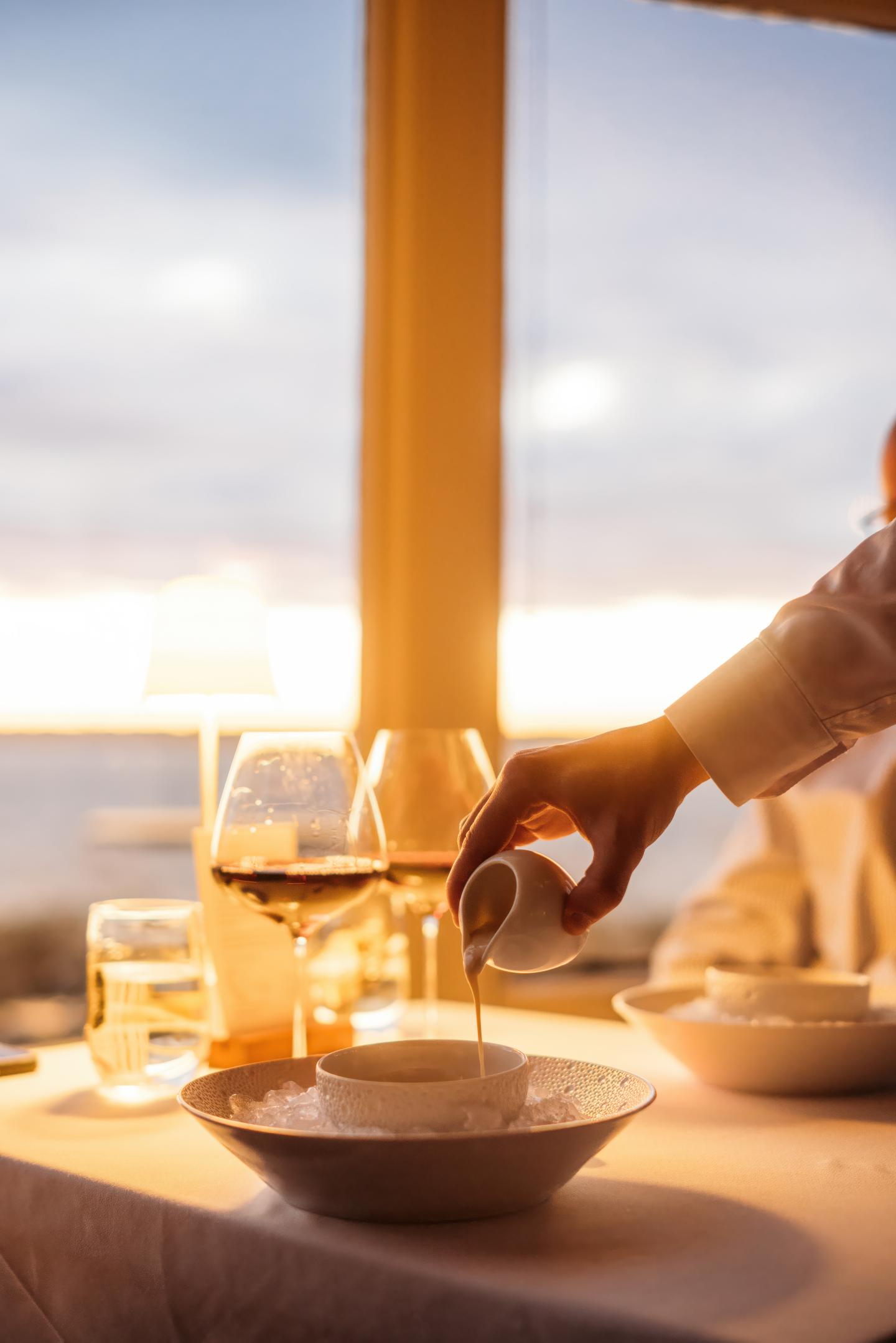 Hand pouring sauce into a bowl on a sunlit dining table, with wine glasses nearby.