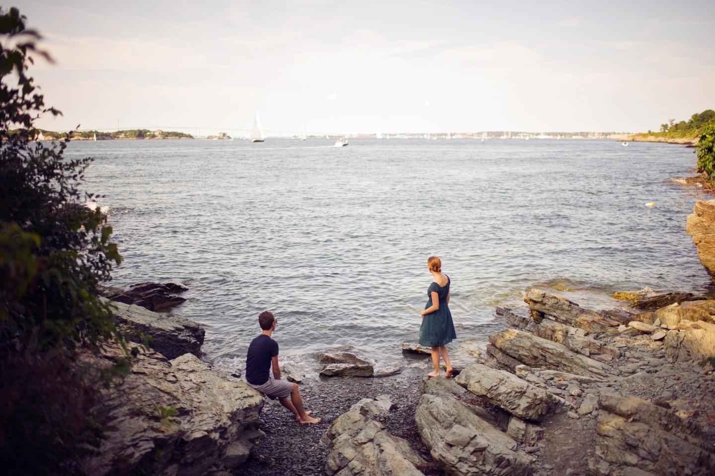 Two people on rocky shore overlooking a calm sea.