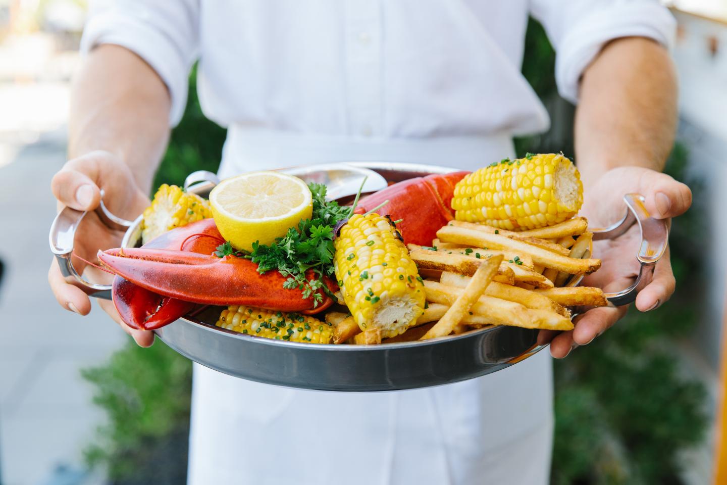 Lobster, corn, fries, and lemon on a tray held by a person in a white shirt.