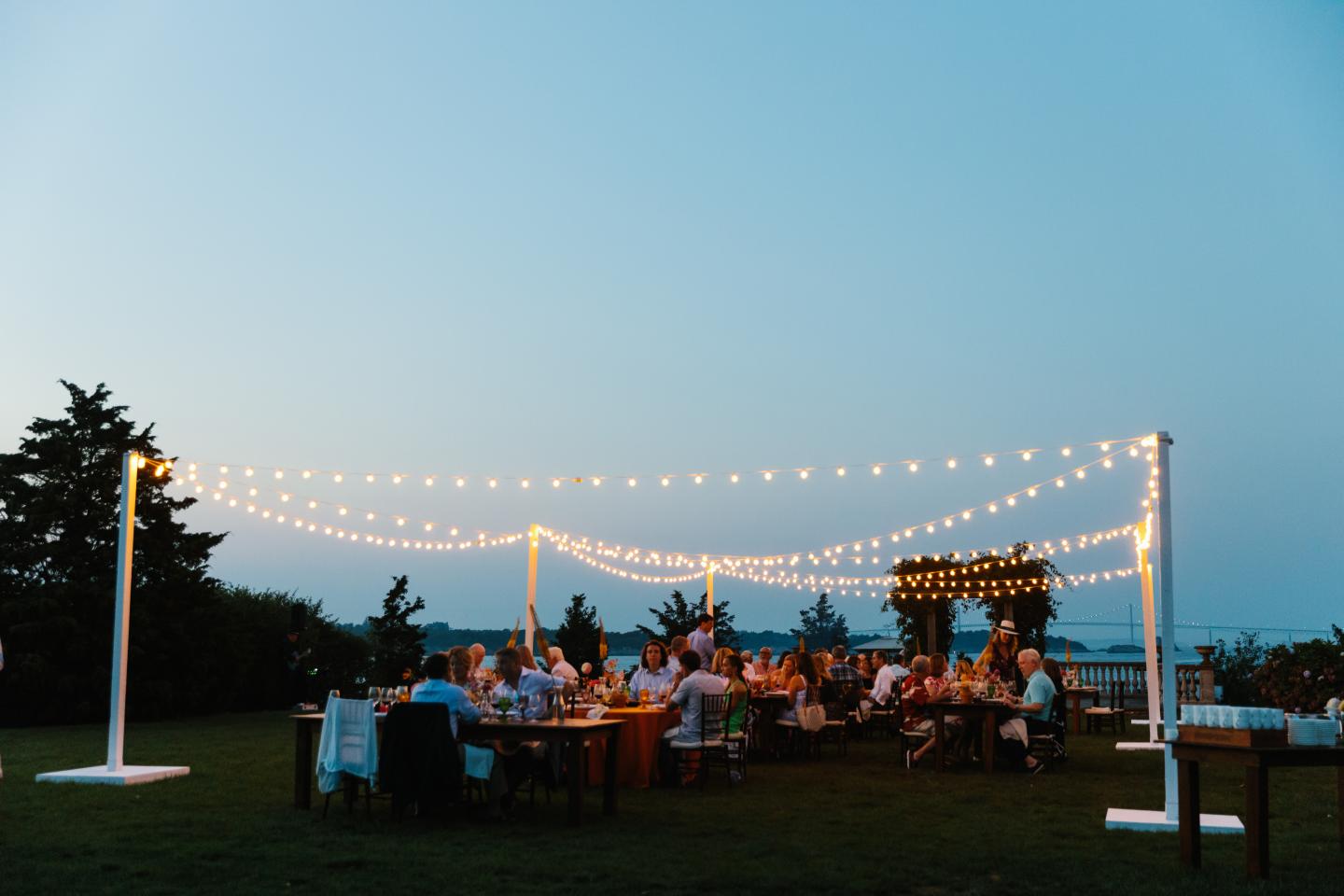 Outdoor evening gathering with string lights and guests at tables.