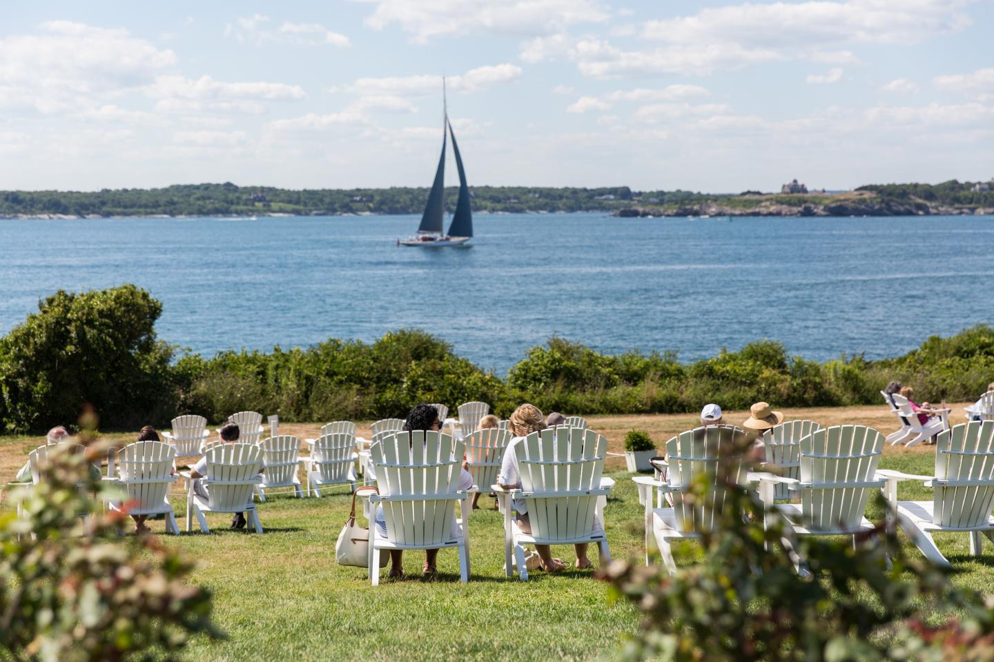 White chairs on a lawn overlooking a calm sea with a sailboat.