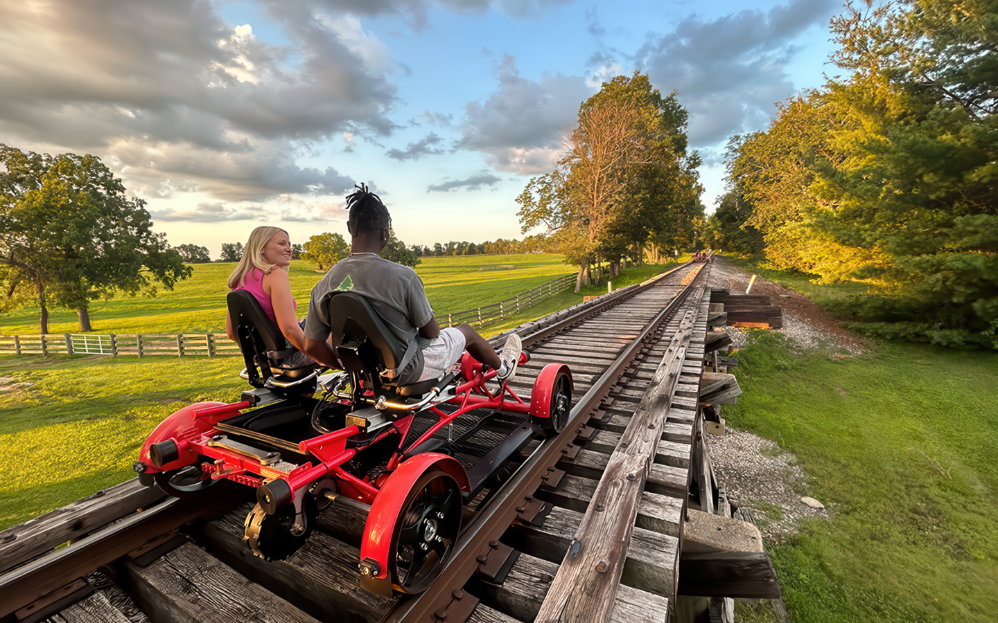 Two people ride a railbike on a wooden track through a green countryside, under a blue sky.