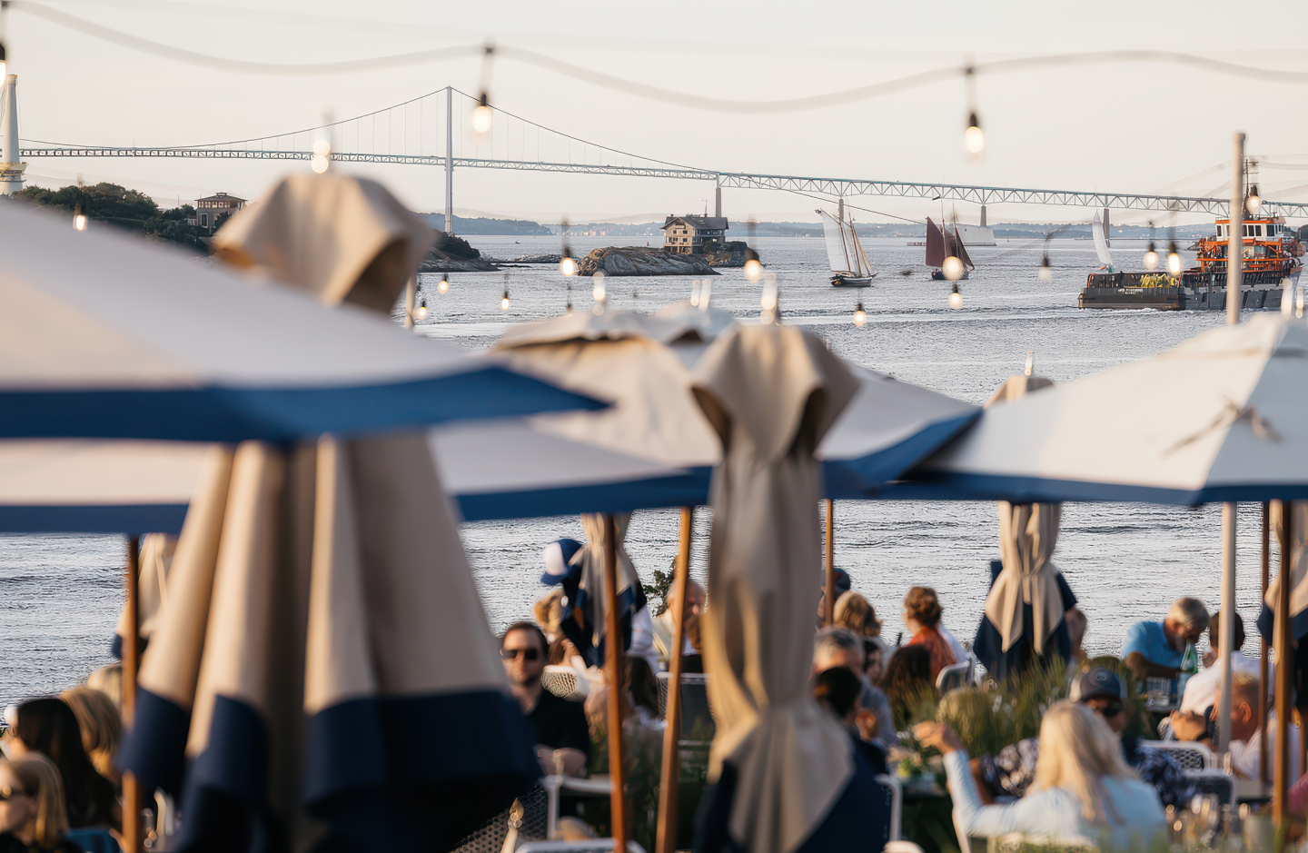 Outdoor café by water, closed umbrellas, bridge, and boats in background.