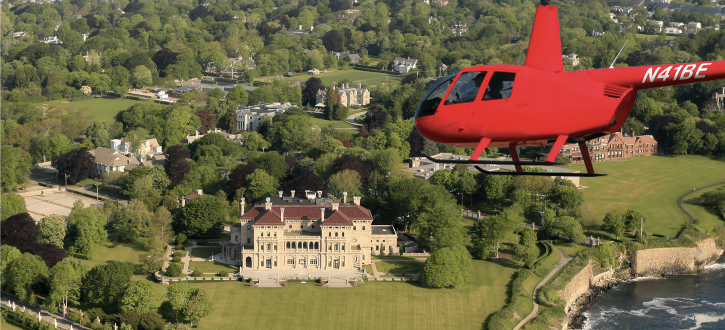 Red helicopter flies over lush estate with mansion below.