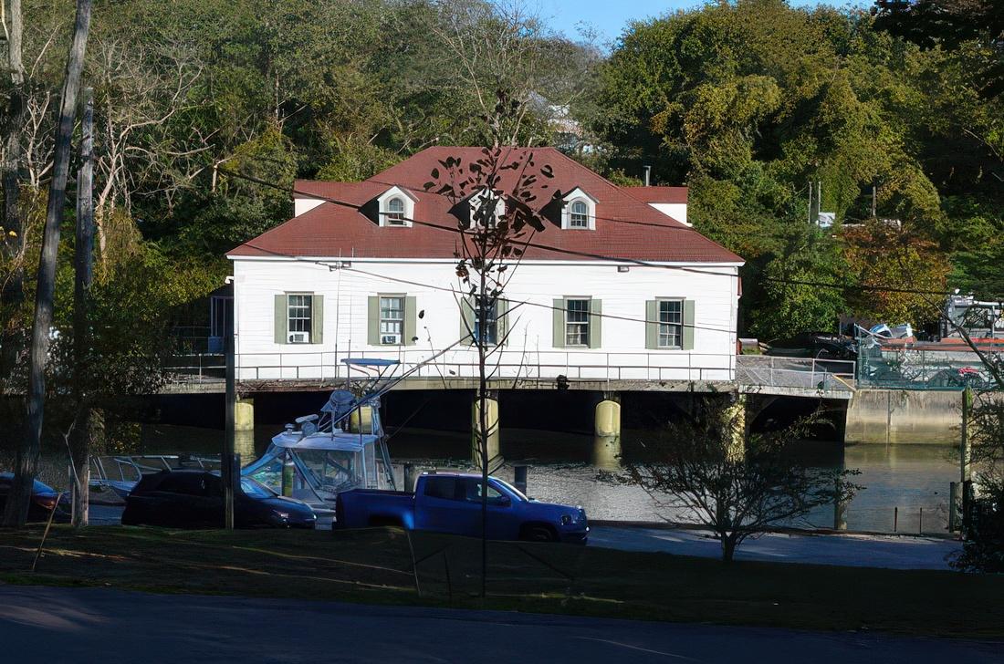 Historic boathouse by a river, surrounded by trees and parked cars in the foreground.