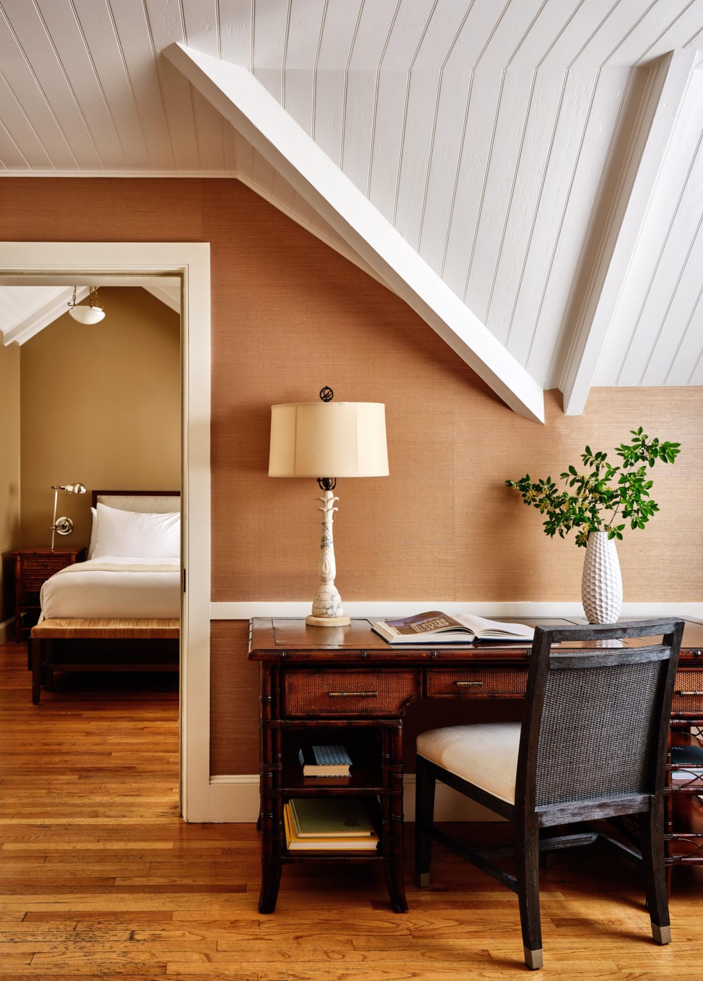 Cozy attic room with desk, chair, lamp, and plant. Bed visible through doorway.