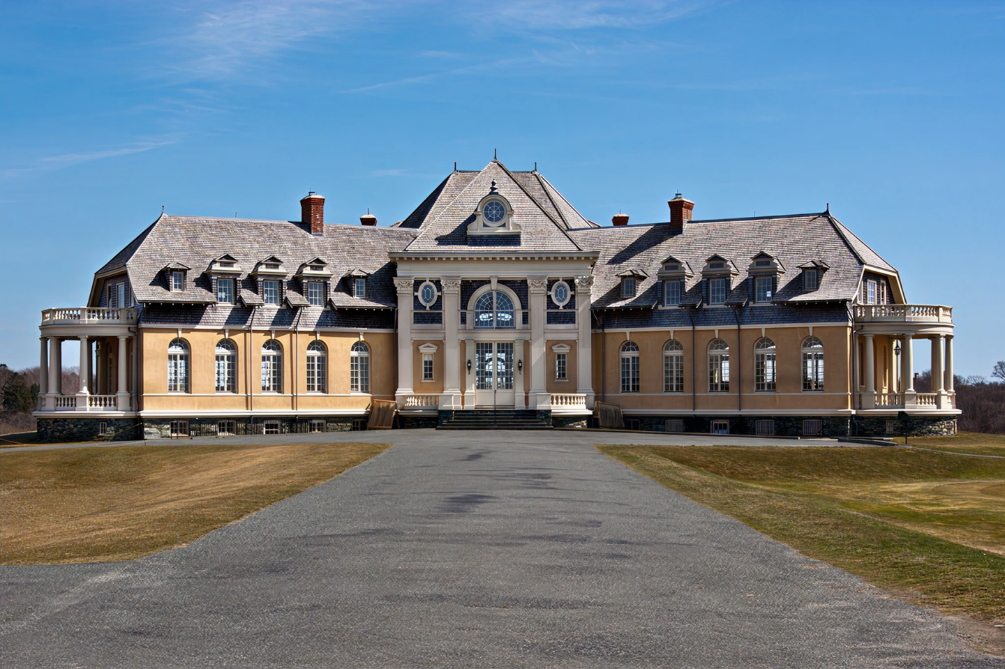 Grand mansion with a long driveway, set against a clear blue sky.