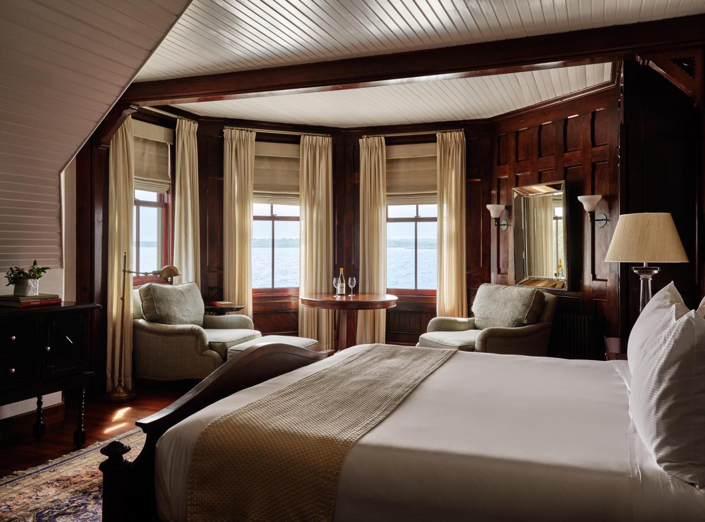 Cozy bedroom with plush bed, armchairs, and bay windows overlooking the water.
