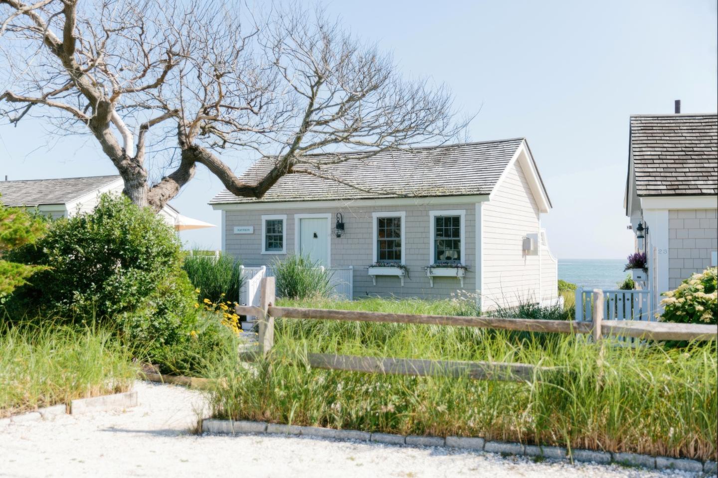 Cozy cottage with a white fence, surrounded by greenery and a view of the ocean.