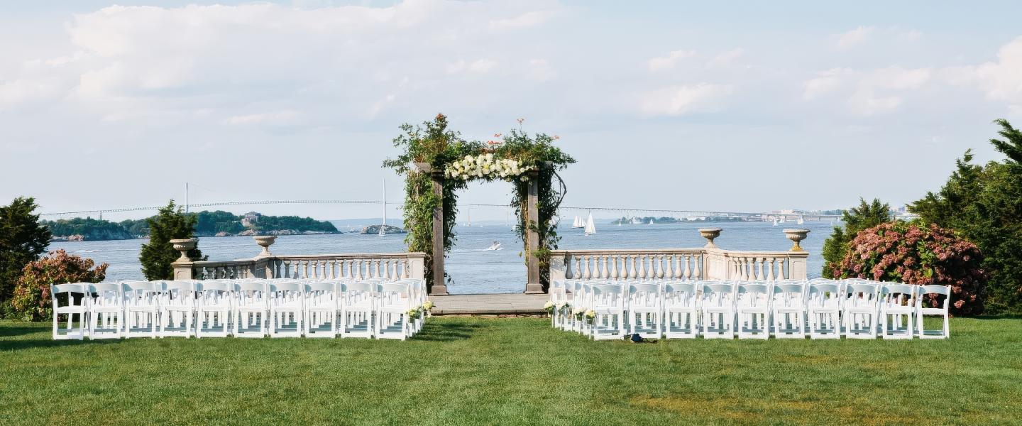 Outdoor wedding setup with white chairs and floral arch by the water.