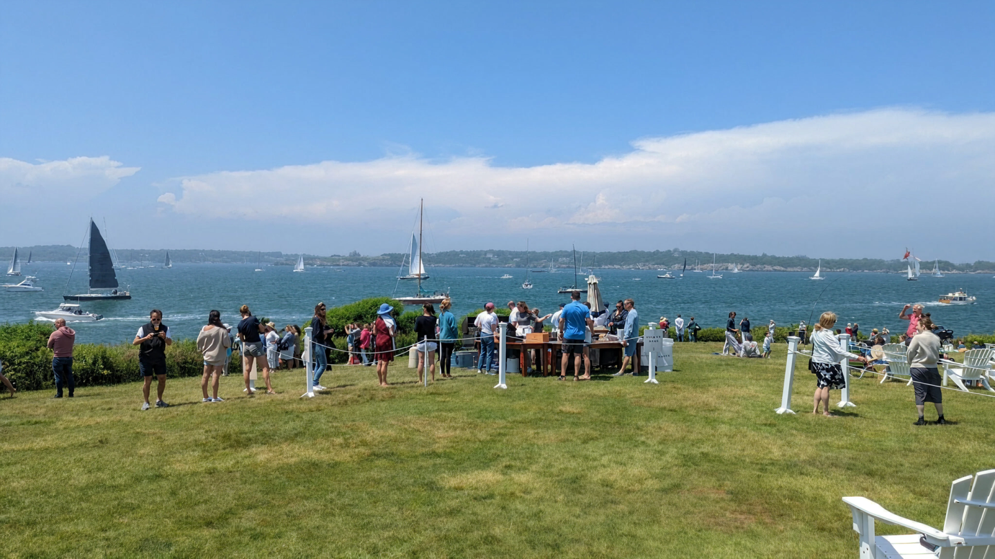 People gathered on a grassy shore, sailboats on blue water in the background.