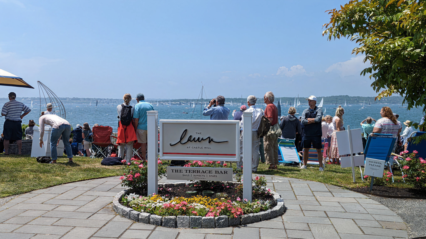 People gather by the waterfront watching sailboats, with a sign and flowers in the foreground.