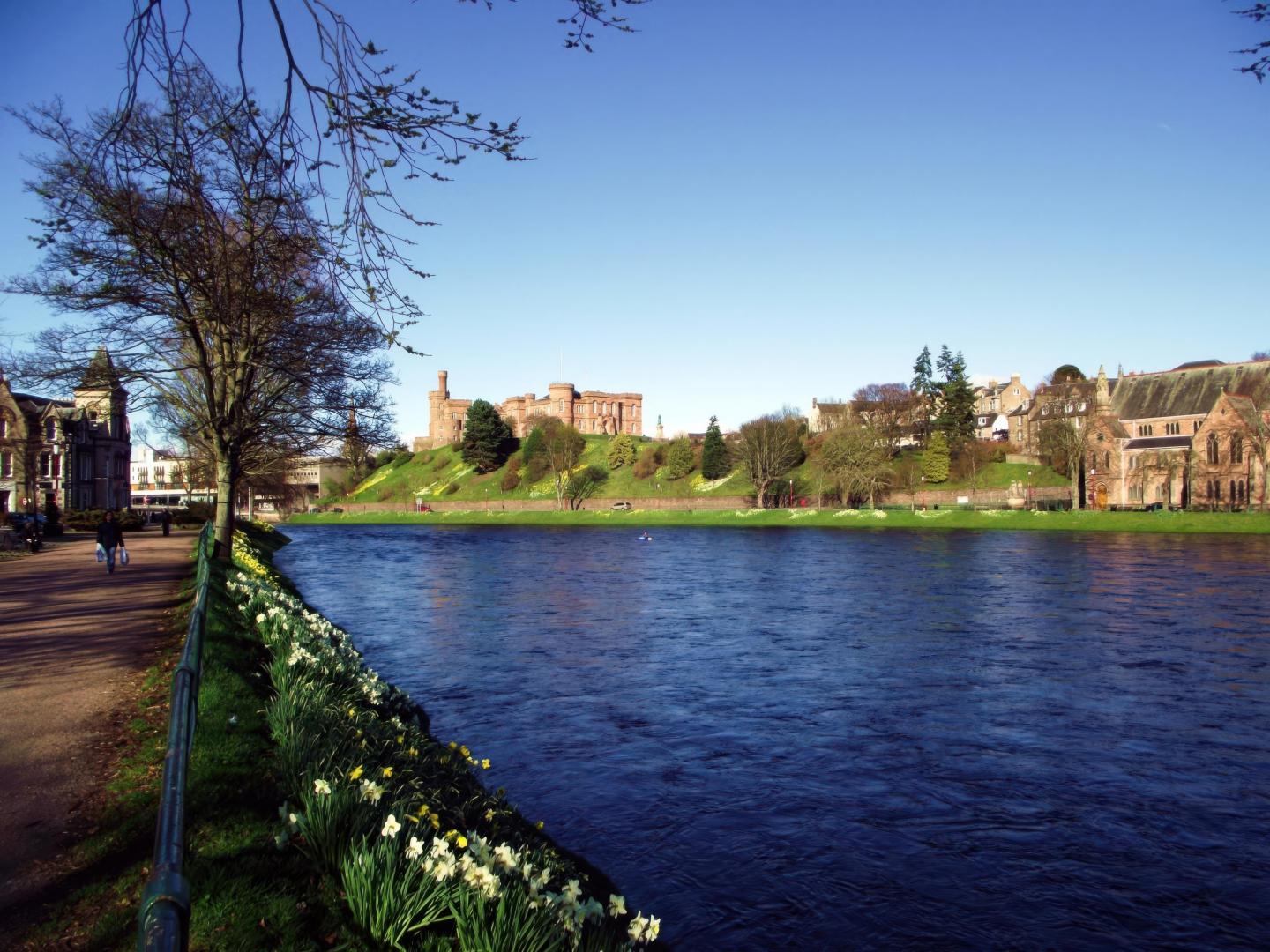 River lined with trees and flowers, buildings in the background under clear blue sky.