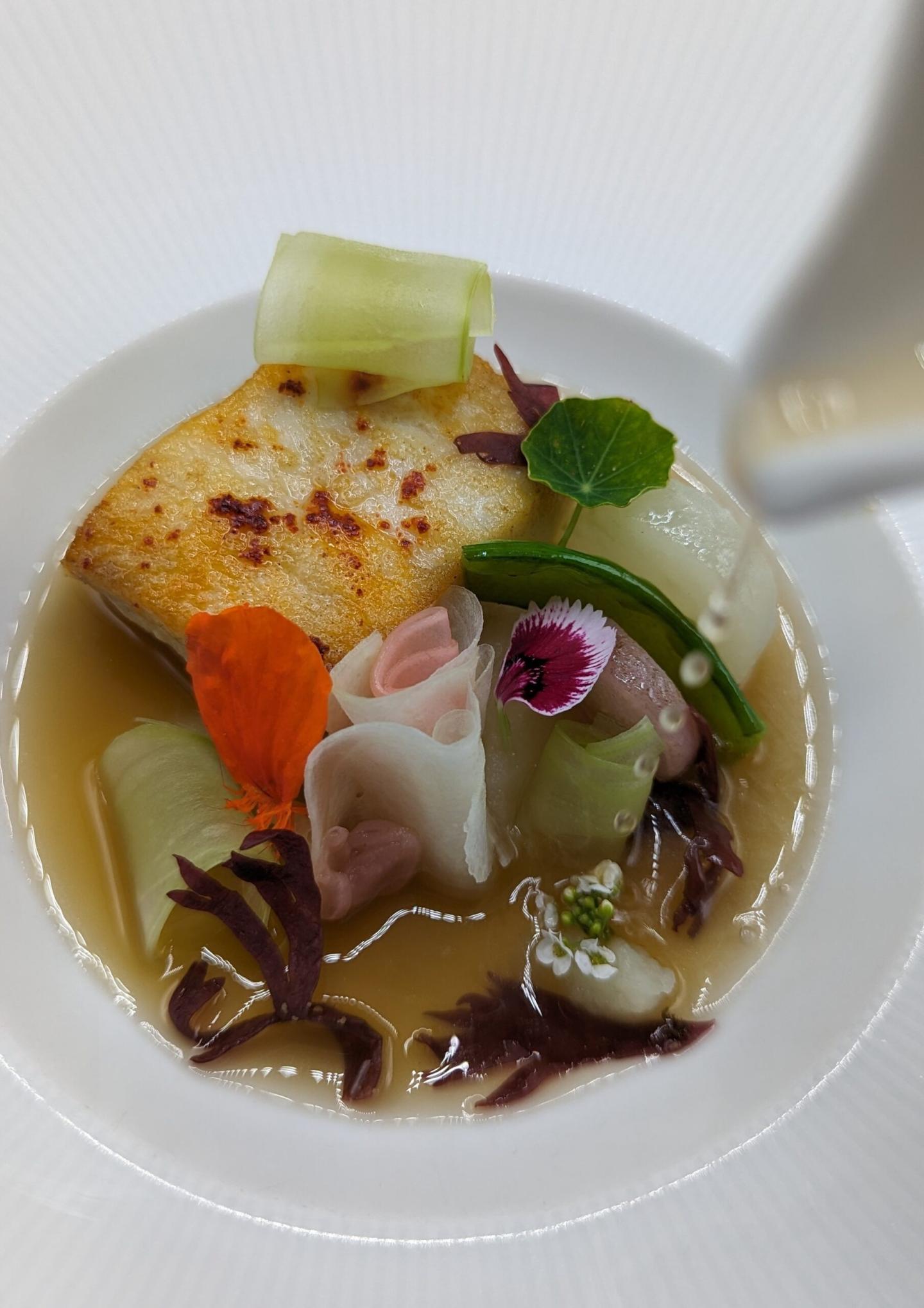 A dish with grilled fish, flowers, and greens in broth.