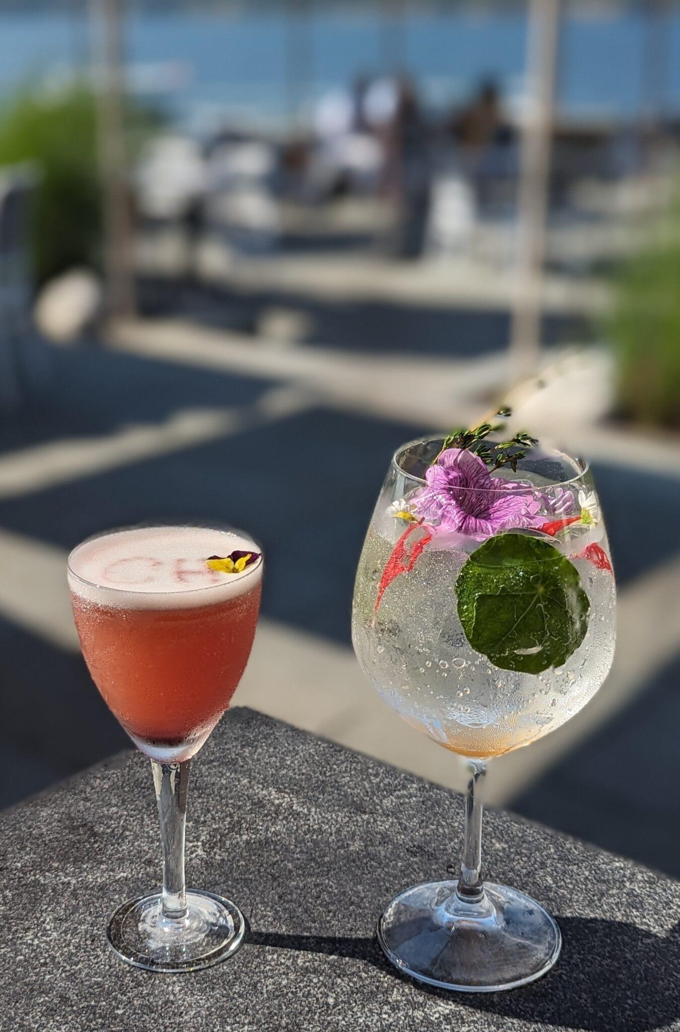 Two cocktails on a table; one pink, one clear with lemon and flowers.