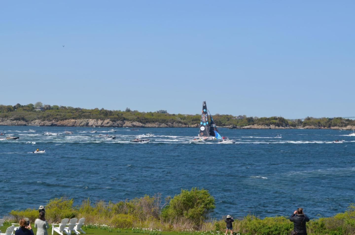 Sailboat race on a blue ocean near a grassy shore under a clear sky.