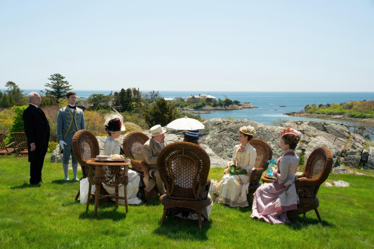 People in vintage attire sit on wicker chairs by the seaside.