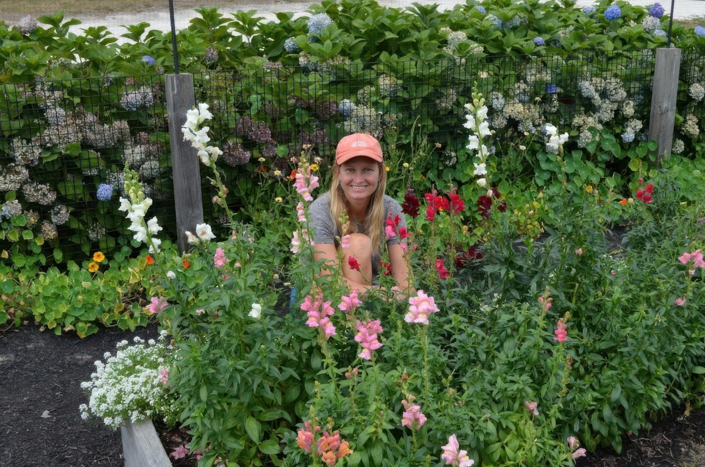Woman in a garden surrounded by colorful flowers, wearing an orange cap and smiling.