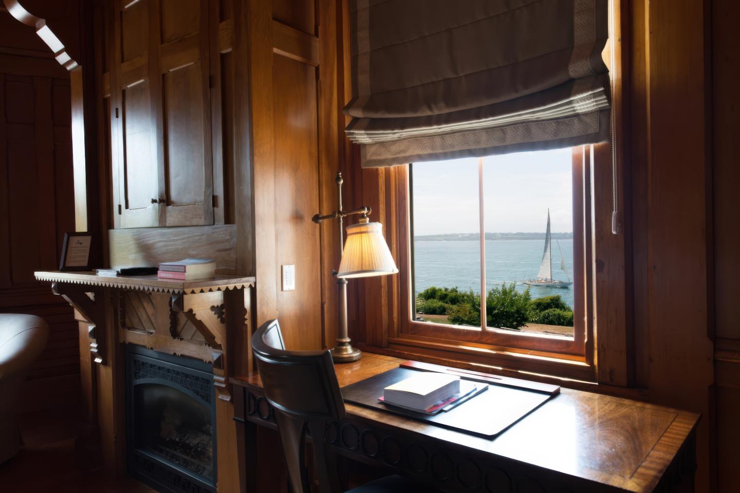 Cozy wooden study with lamp, overlooking ocean and sailboat through window.