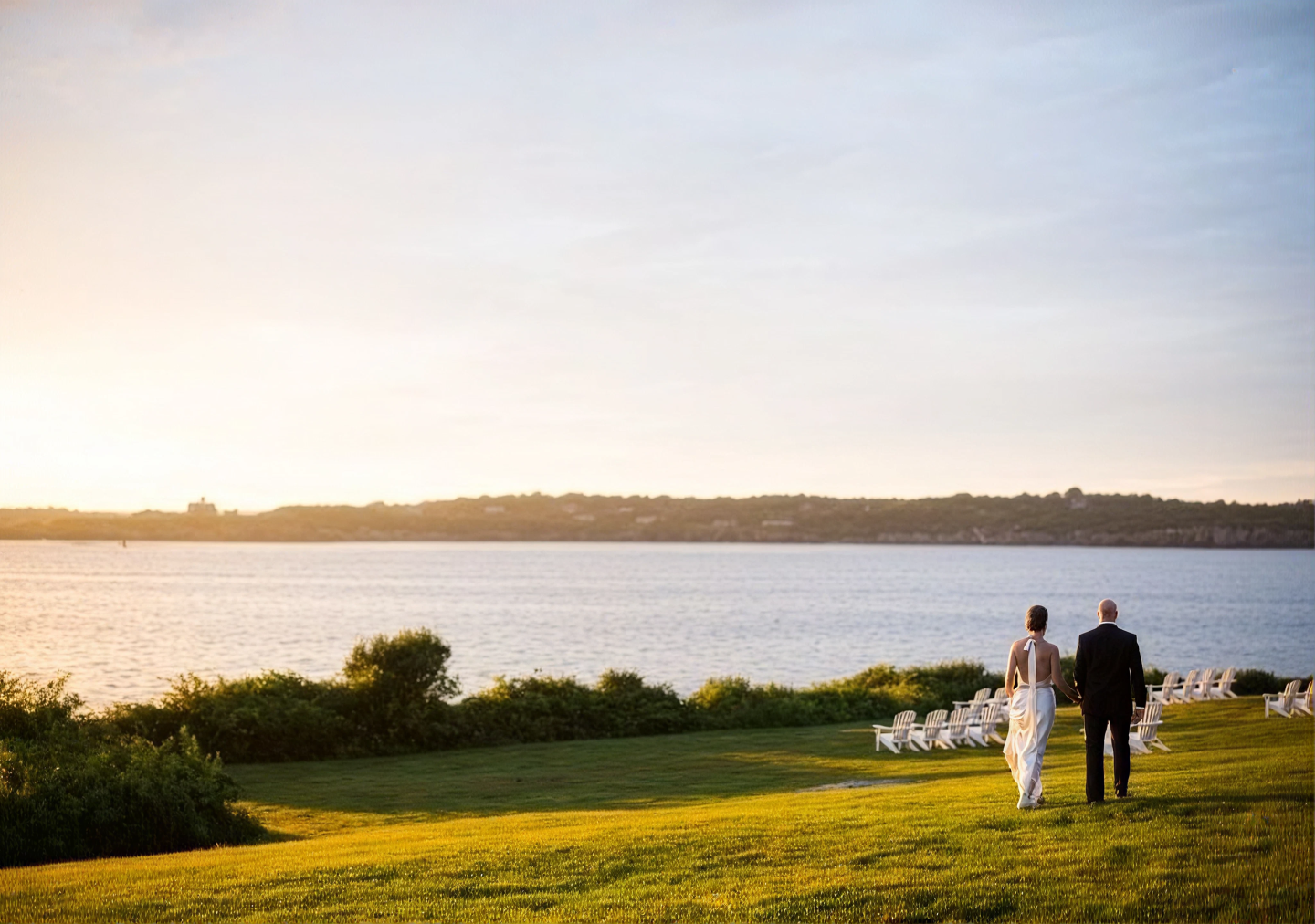 Bride and groom walking on a grassy hill by a lake at sunset.