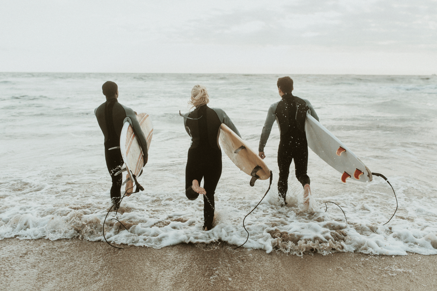 Three surfers in wetsuits walk into the ocean with surfboards.