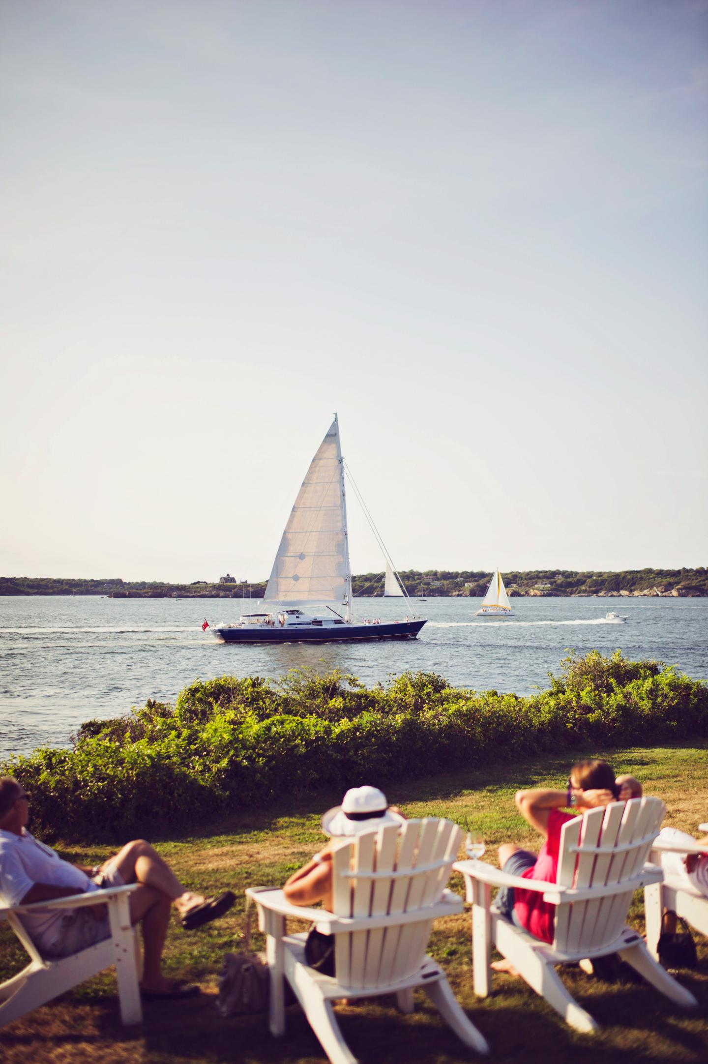 Sailboats on a calm lake, people relaxing in chairs on the grassy shore.
