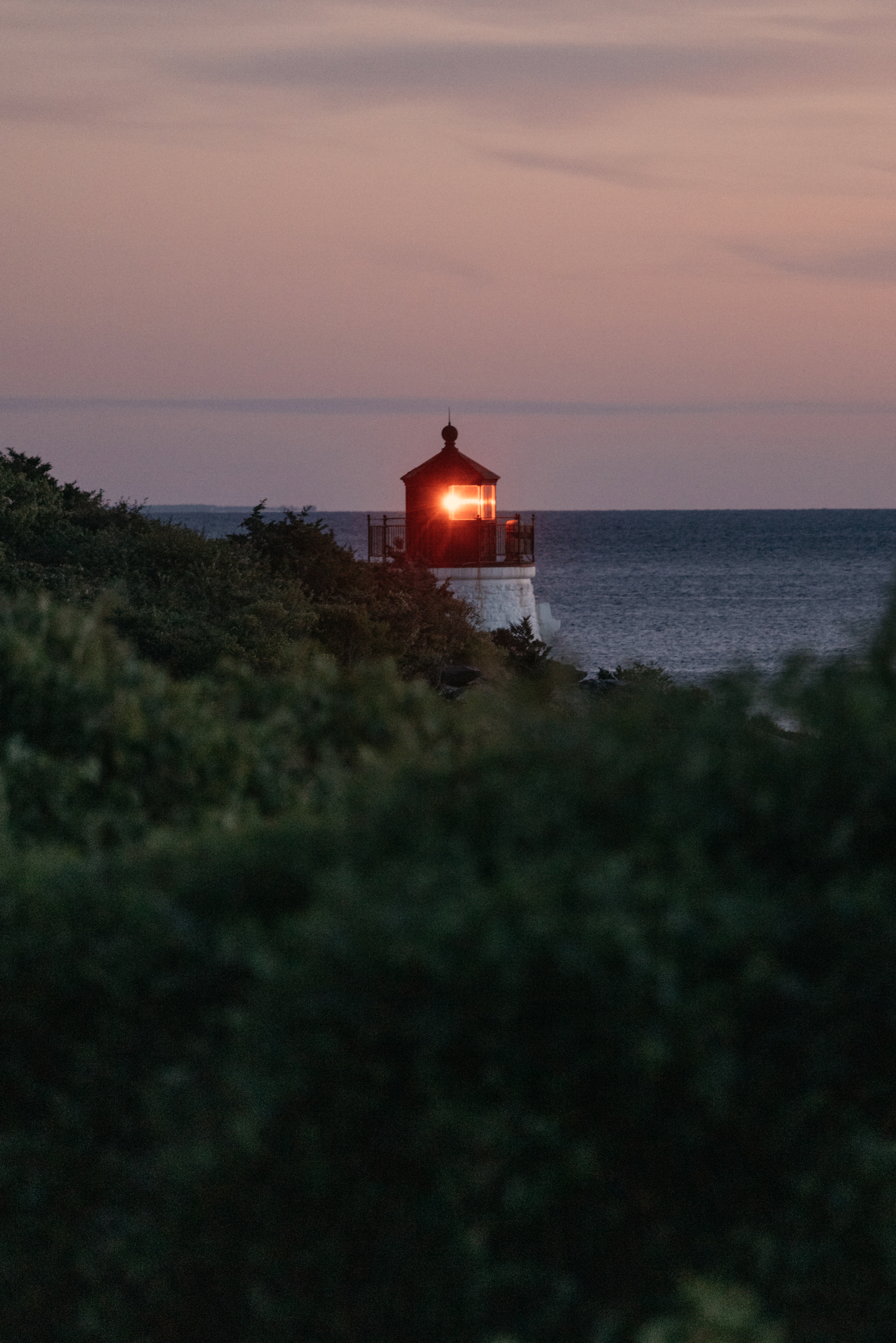 Lighthouse glowing at dusk, surrounded by greenery and overlooking the sea.