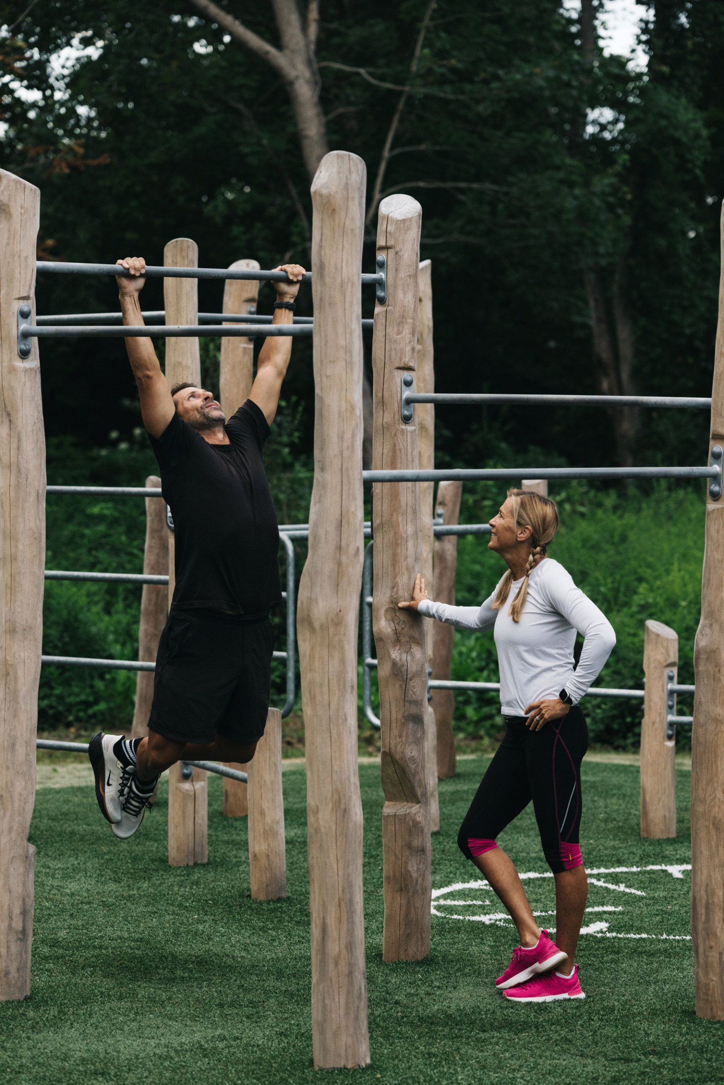 Man doing pull-ups, woman watching in outdoor gym, surrounded by trees.