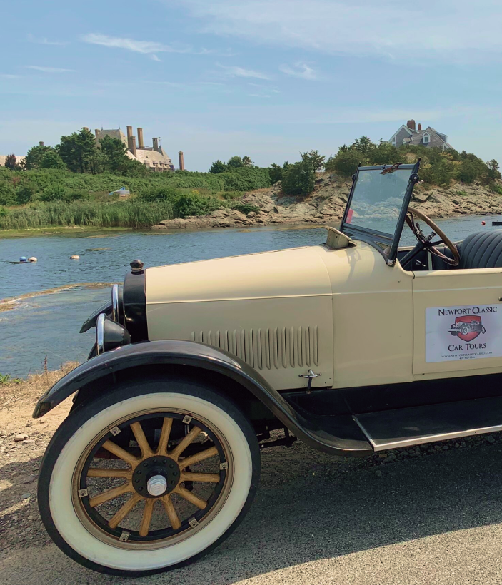Vintage car parked by a lake with ruins in the background.