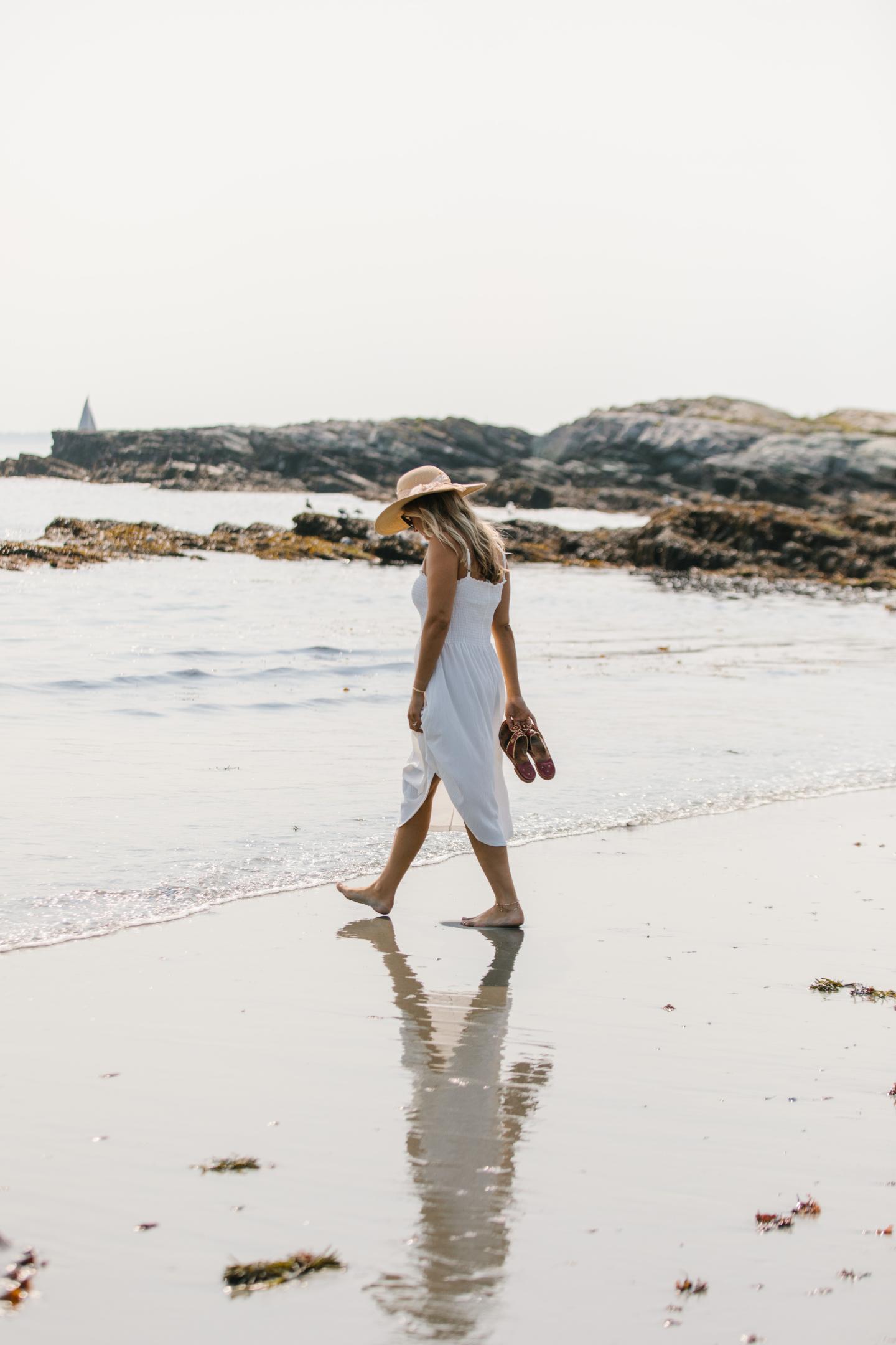 Woman in white dress and hat walking on a beach, holding sandals.