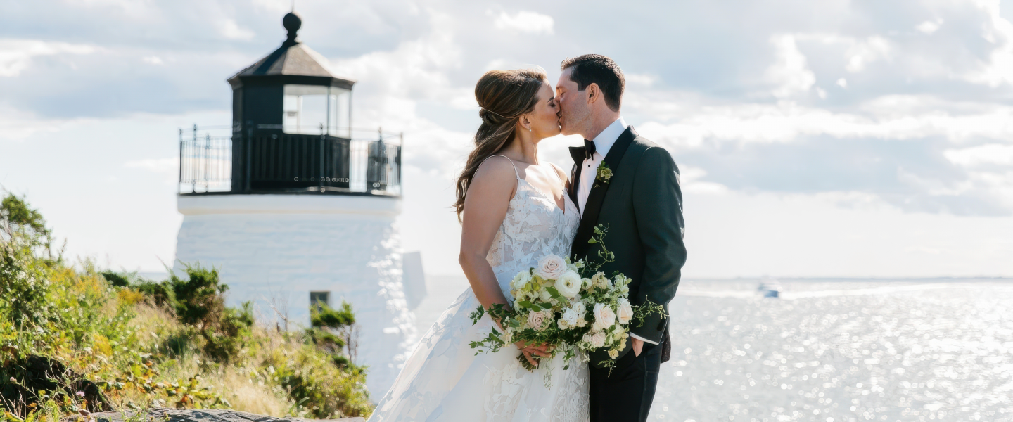 Bride and groom kissing by a lighthouse, with ocean in the background.