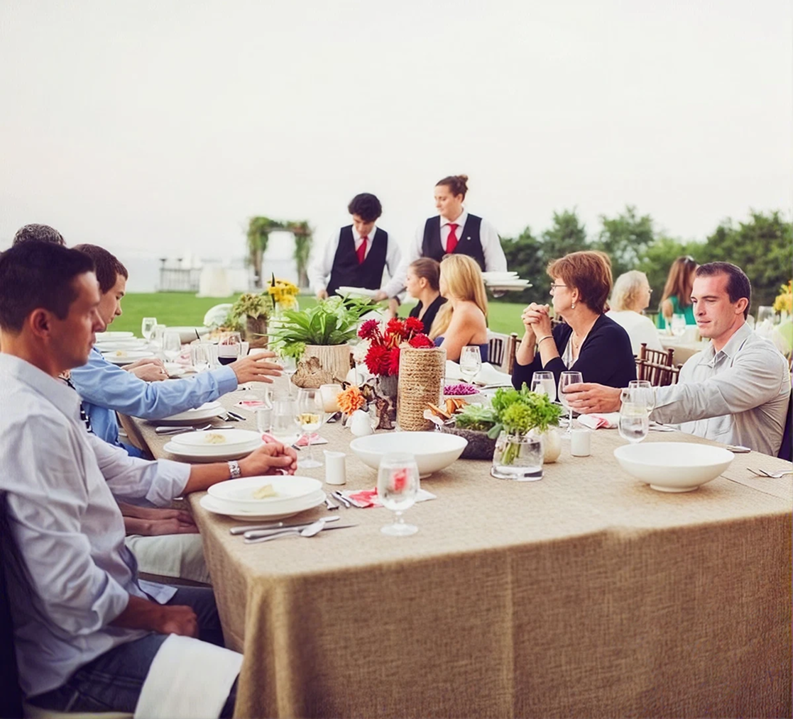 Outdoor dinner party with guests seated around a long table, servers standing nearby.